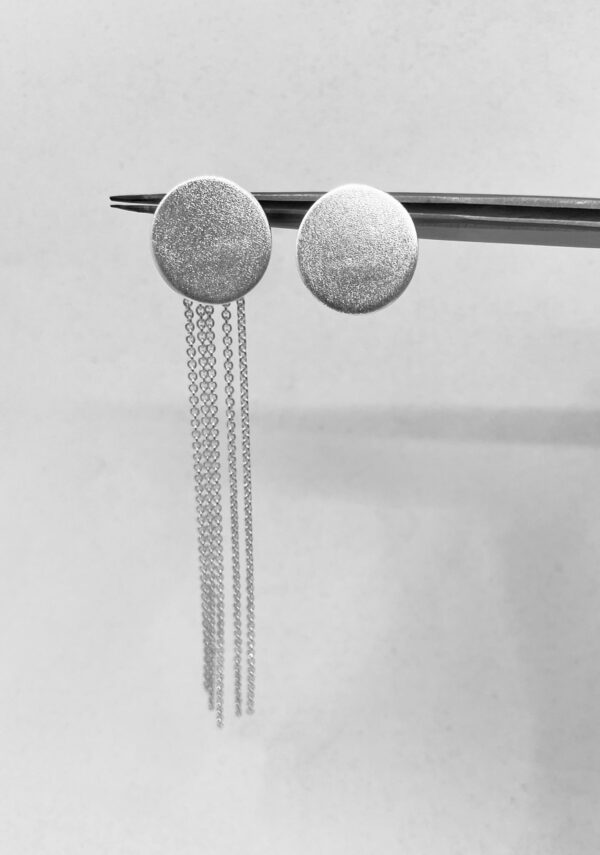 A pair of silver earrings shaped as matte coins, one with hanging thin silver chains and one without chains